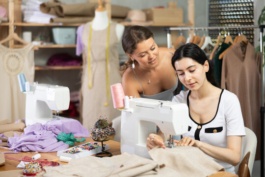 Dedicated young female tailor concentrating on garment stitching at sewing machine, receiving support and advice from teammate in well-equipped clothing atelier