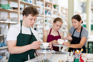 Fototapeta premium Young woman teacher in apron teaches teenage boy and girl students to make ceramic cup in ceramic workshop