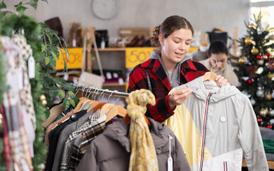Obraz premium Young woman standing choosing a down jacket in a Christmas shop. Buyer at the sale of winter outerwear before the new year