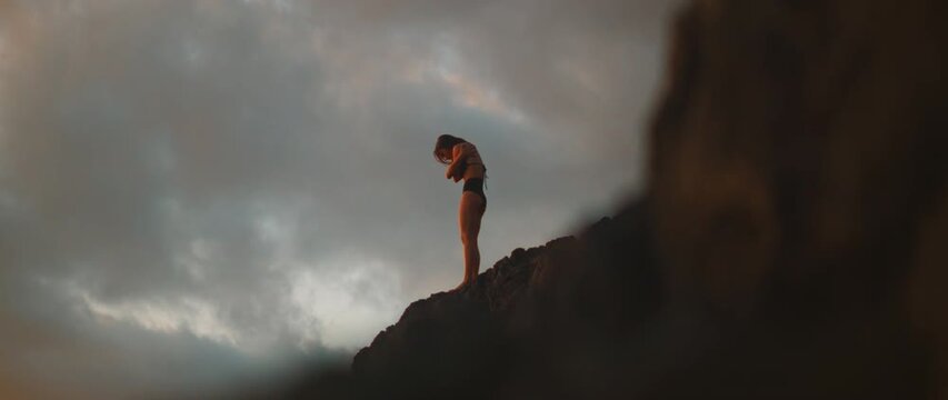 Woman dance hugging herself with both hands in slow movement on rocky peak under moody evening sky