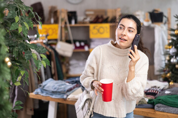 Armenian woman is talking on mobile phone and holding a cup of coffee to take away, she is standing in a clothing store near racks and counters. Shopper in store with Christmas tree background