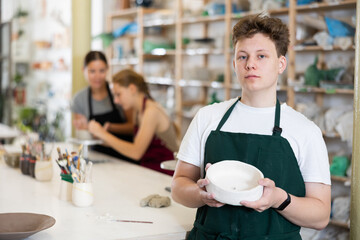 Satisfied teenager guy shows cup created by himself in a clay workshop