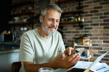 Mature man smiling using smartphone and laptop in home kitchen
