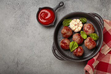 Fried meatballs in a cast iron pan, surrounded by basil, aromatic garlic, and tomato sauce on gray background, healthy home cooking, copy space