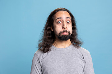 Playful young man with a long hair and beard making a goofy face in the studio. Carefree guy radiates humor and joy, capturing a lighthearted moment full of laughter and casual personality.