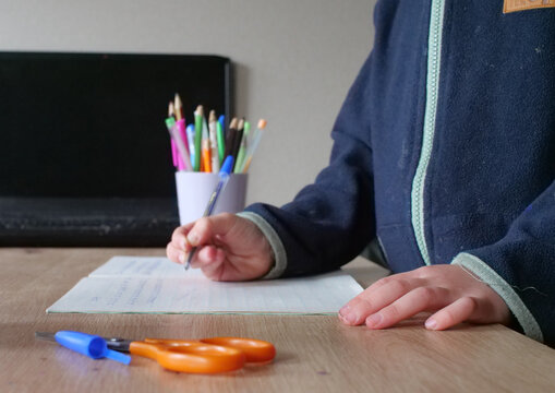 Kid writing at desk with laptop - close-up of a child's hand writing on paper with a pen at a desk with a laptop