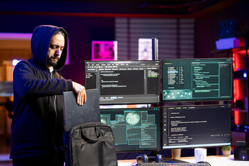 Hacker packing laptop after finishing coding stealth malware to evade security. Evil man putting notebook in backpack after finishing work on script hiding malicious code from detection systems