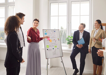 Confident smiling woman presenting ideas on whiteboard to group of professionals in meeting room. Team of business people listening attentively, engaging in discussion, brainstorming during workshop.