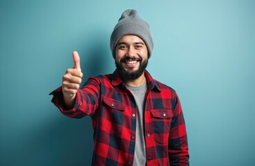 Happy man with beard, mustache wears grey beanie hat, red plaid flannel shirt. Smiles, gives thumbs up gesture, looks confident, positive. Guy shows agreement, success, satisfaction, approval. Adult