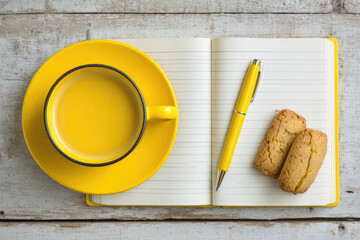Yellow cup filled with tea, matching pen, and two cookies on open lined notebook, arranged on rustic wooden desk, creating cozy and inviting workspace flat lay scene
