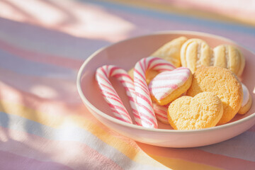 Heart shaped cookies, striped candy canes, and pastel sweets arranged in pink bowl on soft striped fabric create cheerful holiday still life with warm, inviting atmosphere