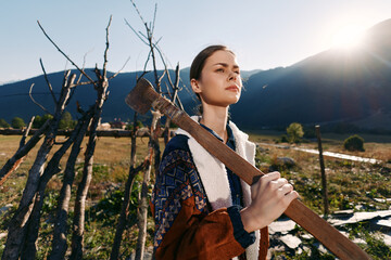 Naklejka premium Woman in countryside field holding an axe over shoulder by wooden fence, mountains in background and sunlight. Rustic portrait of a young farmer in rural landscape and nature.