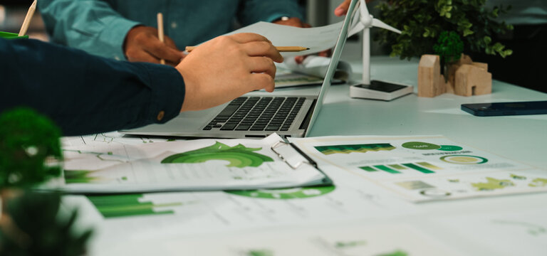 Team members engage in collaborative discussion, analyzing charts and graphs related to sustainability projects, showcasing a modern office with eco-friendly elements and greenery. SACTR
