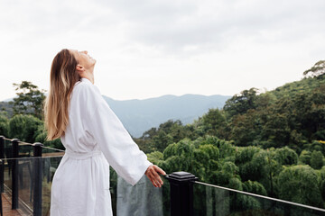 Young woman in white robe relaxing on balcony overlooking lush green mountains