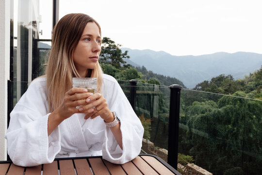 Young woman relaxing with a drink on a balcony overlooking a forested mountain landscape - Powered by Adobe