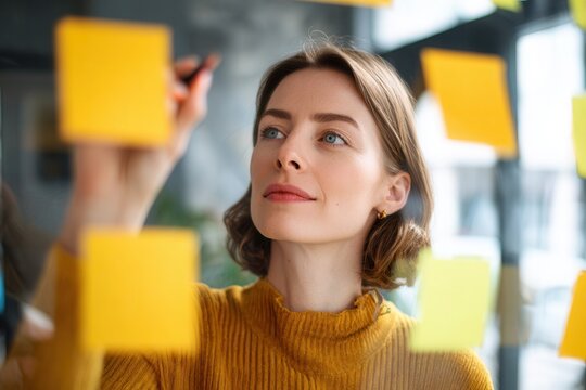 Creative businesswoman brainstorming ideas on glass wall with sticky notes, planning strategy for success in modern office, fostering innovation and collaboration