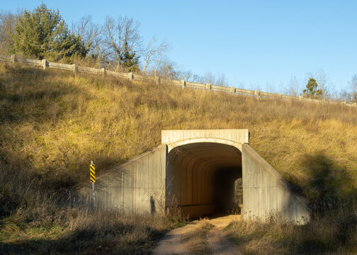 The entrance of a former-railroad tunnel crossing under a rural highway on a sunny autumn day