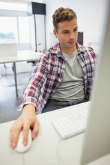 Man in 20s sitting in workspace using computer with keyboard and wired mouse, wearing plaid shirt