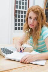 Teenage female studying at wood table with textbook and laptop holding pen wearing turquoise top