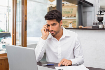 Man 25-35 wearing white shirt, holding smartphone and pointing at papers by laptop in cafe