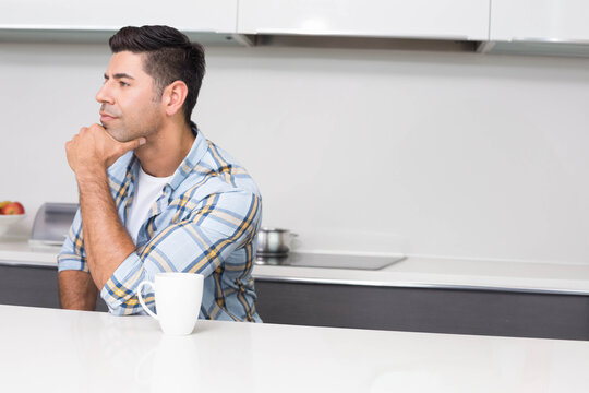 Man sitting at kitchen counter wearing plaid shirt, resting chin, looking toward cooktop with mug