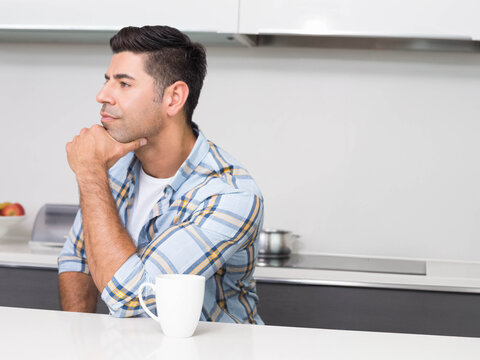 Man sitting at white kitchen countertop wearing plaid shirt, resting chin, holding mug - Powered by Adobe