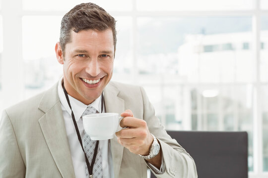 Middle-aged man wearing beige suit and ID lanyard, smiling while holding white coffee cup in office - Powered by Adobe