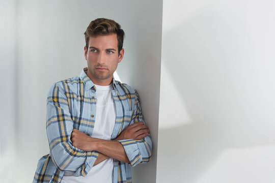 Man leaning against wall in studio, arms crossed, wearing plaid shirt and white T-shirt, copy space