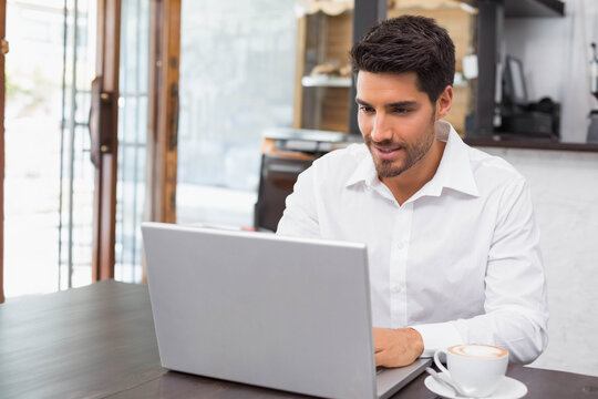 Man wearing white shirt working on silver laptop in coffee shop on wooden table with cappuccino