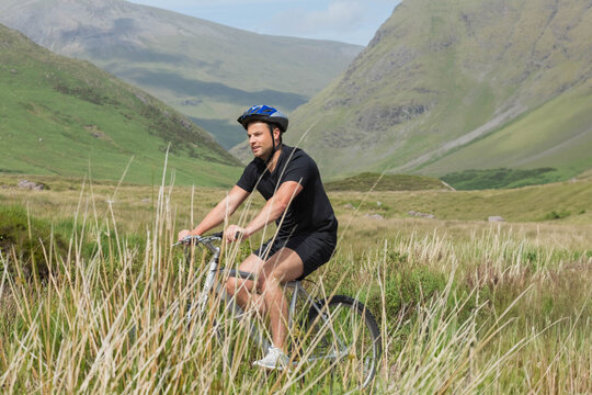Adult male cyclist pedaling bicycle through tall grasses in wide green valley wearing blue helmet - Powered by Adobe
