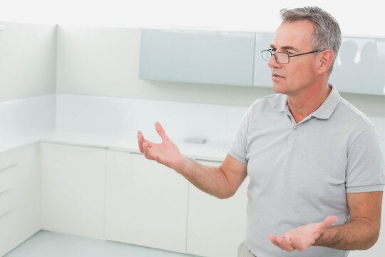 Senior man wearing gray polo and glasses gesturing in white kitchen, phone on counter, copy space