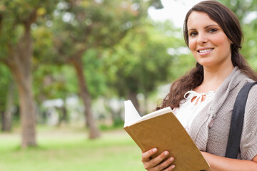 Fototapeta premium Teenage woman standing holding open hardcover book in park wearing cardigan, bag strap, copy space