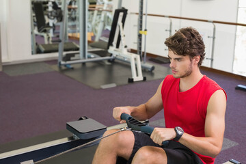 Adult man pulling foam handle on rower at gym wearing red sleeveless shirt wristwatch, copy space