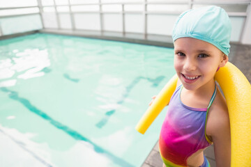 Child standing at pool edge holding yellow noodle in colorful swimsuit, blue swim cap, copy space