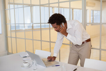 African American man in white shirt leaning over silver laptop holding phone in office, copy space