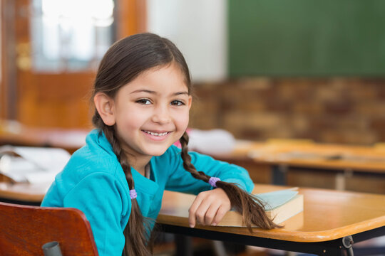 Asian girl child sitting at wooden desk in classroom wearing turquoise top, resting hand on book