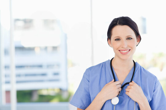 Adult female clinician standing, smiling, in blue scrubs holding stethoscope in clinic, copy space