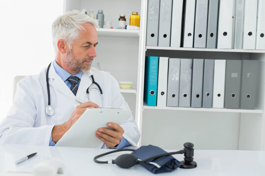 Senior male physician sitting at desk in clinic wearing lab coat, holding stethoscope and clipboard