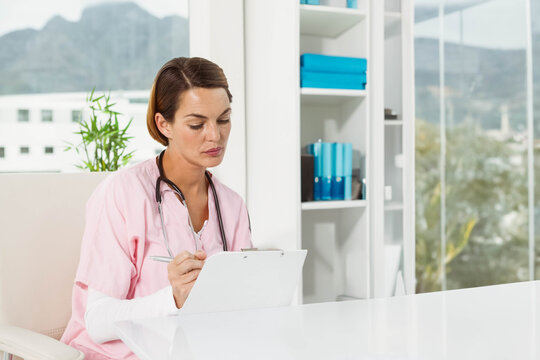 Female in pink scrubs sitting at desk in medical office writing on clipboard wearing stethoscope