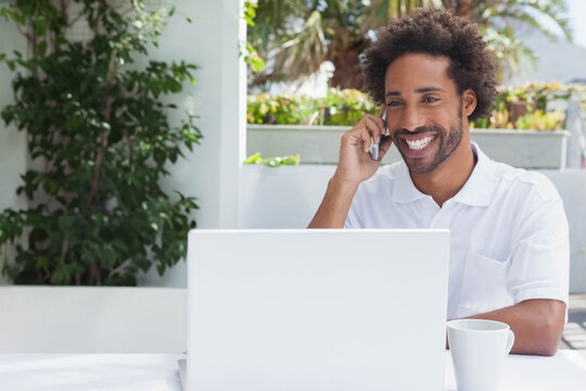 African American man in white polo using laptop while phoning with coffee and planters on terrace