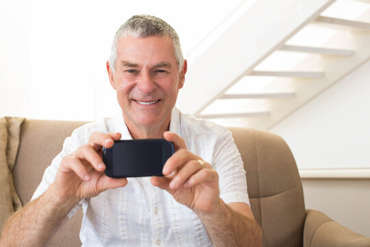 Senior man in living room sitting on armchair holding phone tilted showing wedding ring near stairs
