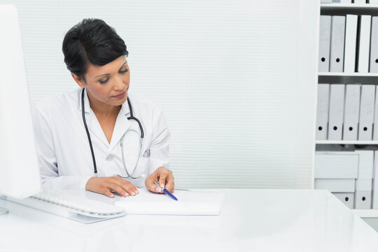 Asian female physician in white lab coat sitting at desk writing with blue pen wearing stethoscope - Powered by Adobe