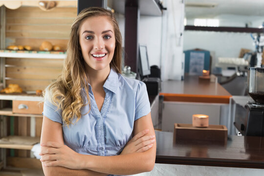 Female barista standing at counter in light blue blouse, showing wooden shelves and coffee grinder