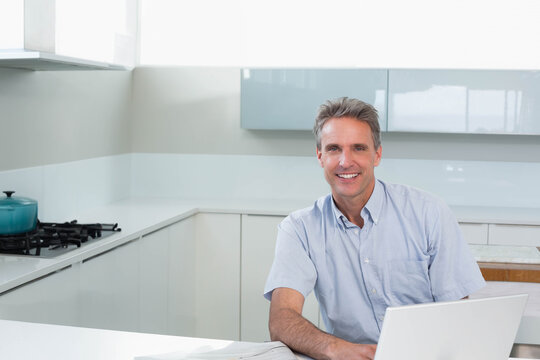 Middle-aged man in blue shirt sitting at kitchen counter using laptop beside teal pot, copy space