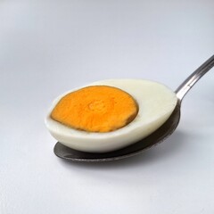 Boiled egg in a spoon. A boiled egg sliced in half is displayed on a metal spoon against a light background. The yolk is a vibrant orange, surrounded by a smooth, white cooked egg white. 