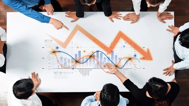 A diverse business team collaborates in a modern conference room, analyzing data trends on a table with graphs and charts, highlighting market fluctuations and strategies. Raster