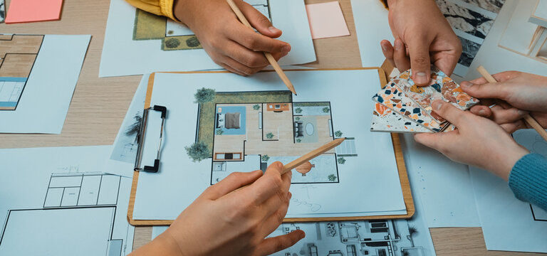 A diverse group of individuals collaborates on design layouts and sketches at a modern workspace. Various materials and tools are arranged on a table, showcasing teamwork and creativity. SACTR - Powered by Adobe