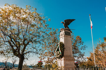 War memorial statue of an eagle atop a stone pedestal with the Italian flag, surrounded by autumn...