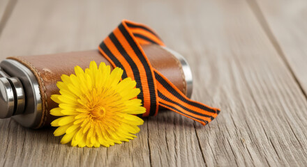 A vibrant yellow dandelion and an old soldier's leather flask with St George's ribbon on rustic wood, symbolizing remembrance.