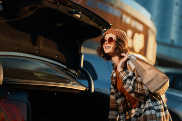 Woman by car trunk with bag and sunglasses wearing beanie and plaid coat, golden hour candid...